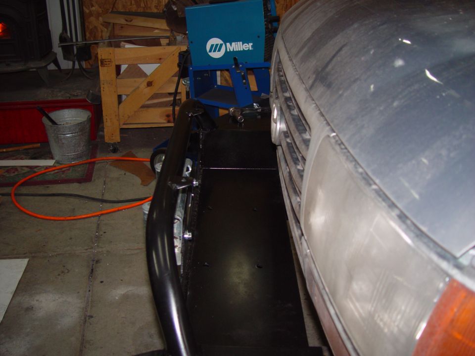 Front corner of a dusty vehicle, showing headlight and grille. A black metal bumper is attached. Miller welding machine in background