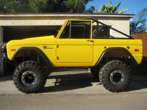 1970 Ford Bronco - A bright yellow 1970 Ford Bronco is viewed from the side, modified with large off-road tires and a black roll cage.