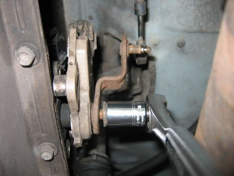 1992 Ford Bronco - Close-up under a vehicle showing a rusty steel bracket and a chrome socket on a ratchet or breaker bar engaging a bolt, with a cast metal c…