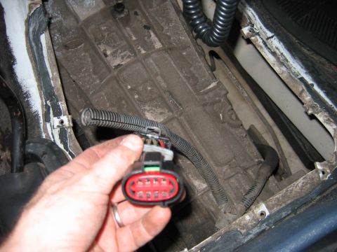 1992 Ford Bronco - A hand holds a black multi-pin electrical connector with a red locking body and green tab, attached to a corrugated wiring loom, in front o…