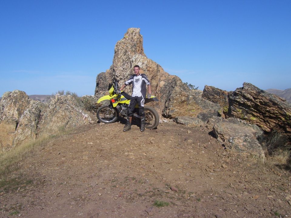 Man in riding gear stands with yellow dirt bike on a rocky hill under a clear blue sky. Supermotors.net logo in corner