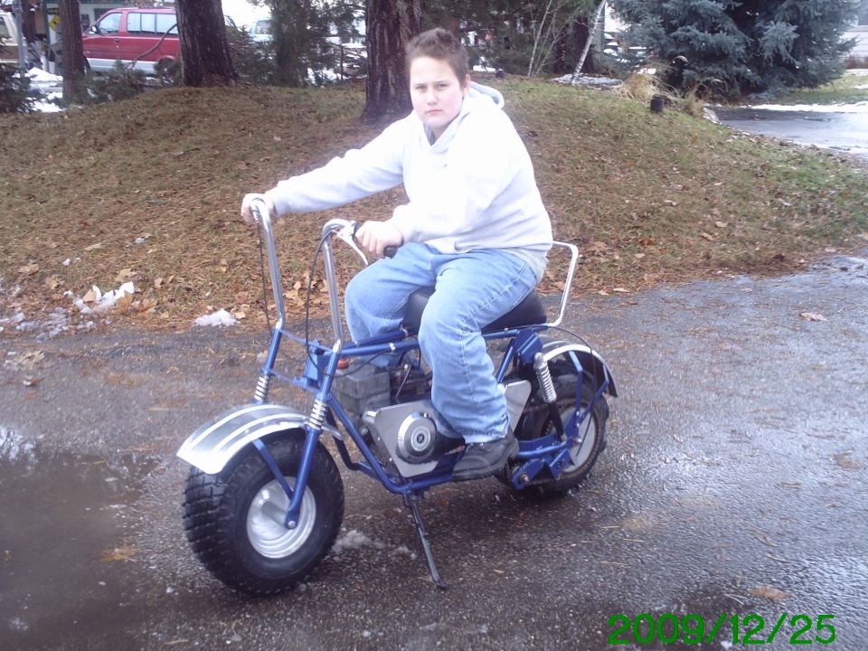 Youth sitting on a blue mini bike with silver fender on a wet driveway