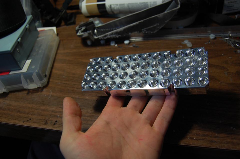 Hand holding chrome multi-cup lamp reflector panel over a cluttered workbench