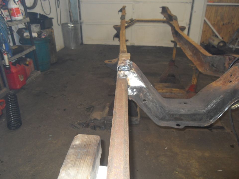 rusted vehicle frame on jack stands in a garage, with a spring and gas cans nearby