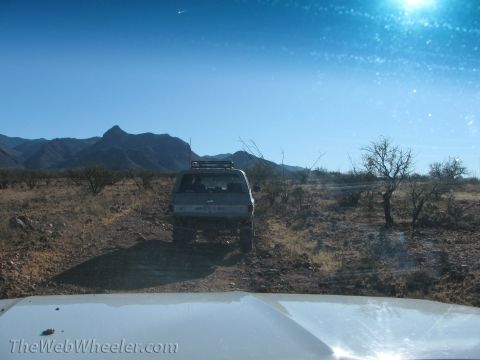 1996 Ford C-350 (Centurion Conversions) - Two vehicles driving on a dirt track through a desert landscape with mountains.