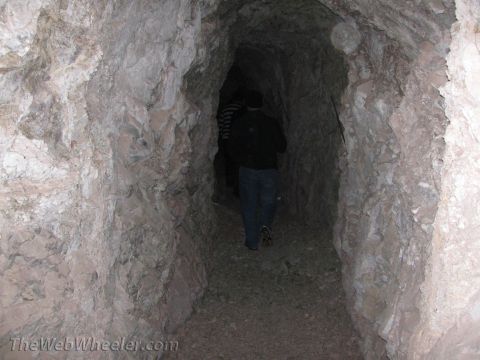 1996 Ford C-350 (Centurion Conversions) - A dark cave tunnel with rough rock walls and a dirt floor, where two people are walking into the darkness.
