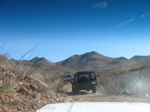 1996 Ford C-350 (Centurion Conversions) - Two SUVs navigate a dusty desert trail under a bright blue sky, with arid mountains in the background.