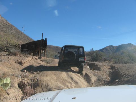 1996 Ford C-350 (Centurion Conversions) - A black SUV navigates a dusty trail with a wooden structure on the left and mountains in the distance.