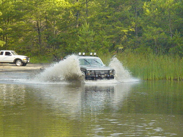 1980 Ford Bronco splashing through water, white pickup truck parked on shore