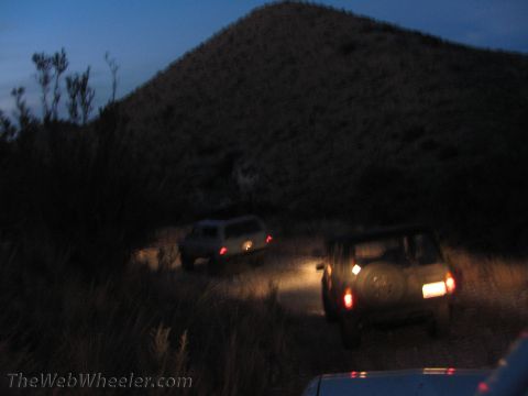 1996 Ford C-350 (Centurion Conversions) - Two SUVs navigate a dirt road at twilight, their headlights cutting through the dim light.