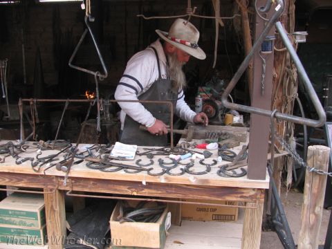1996 Ford C-350 (Centurion Conversions) - A blacksmith crafts metalwork, displaying finished horseshoes and tools on a wooden table.