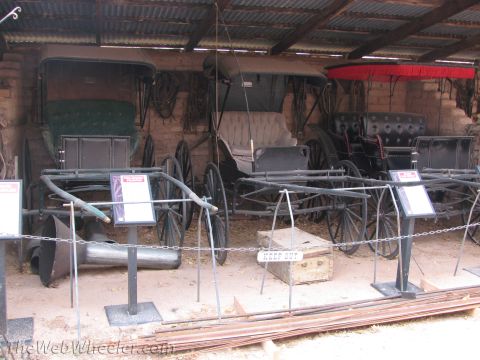 1996 Ford C-350 (Centurion Conversions) - A collection of antique horse-drawn carriages are displayed indoors, with informational signs in front of them.