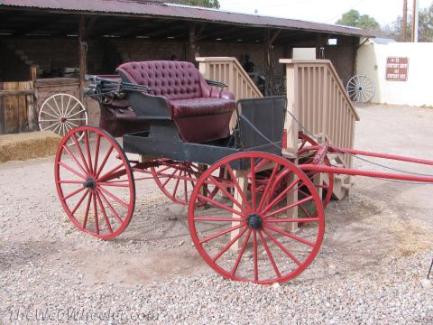1996 Ford C-350 (Centurion Conversions) - A vintage horse-drawn carriage with striking red wheels and a plush burgundy interior sits outside a rustic building.