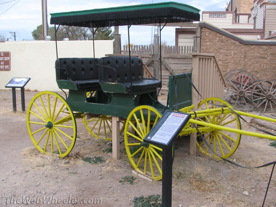 Green and yellow horse-drawn carriage with black tufted seats and a fringed canopy. Signage is visible