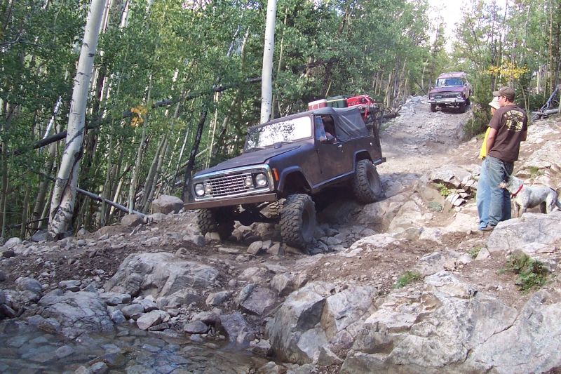 Several off-road vehicles navigate a steep, rocky trail with trees on the side. A man and dog stand watching