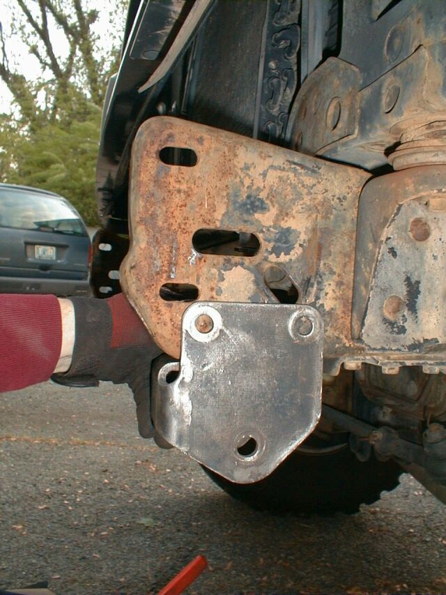 Rusty leaf-spring shackle bracket on a vehicle frame with new bare-metal mount welded below, viewed from underneath