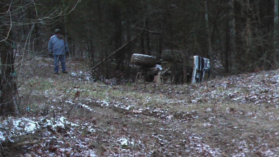Truck rolled on its side off a leaf-covered dirt trail in bare winter woods, man walking toward it