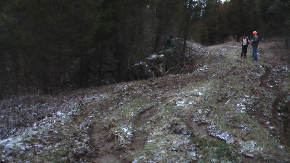 Rutted muddy trail with a rolled light-colored truck at the tree line and onlookers standing nearby