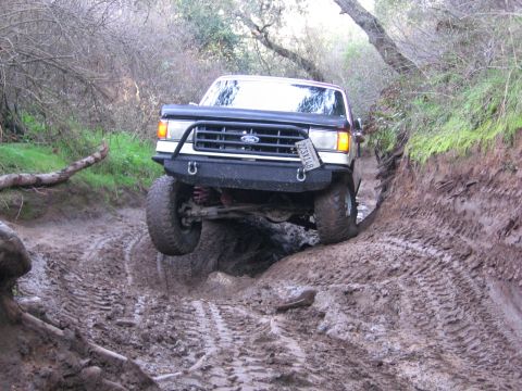 1989 Ford Bronco - A Ford Bronco drives through a muddy off-road trail, with one front wheel lifted.