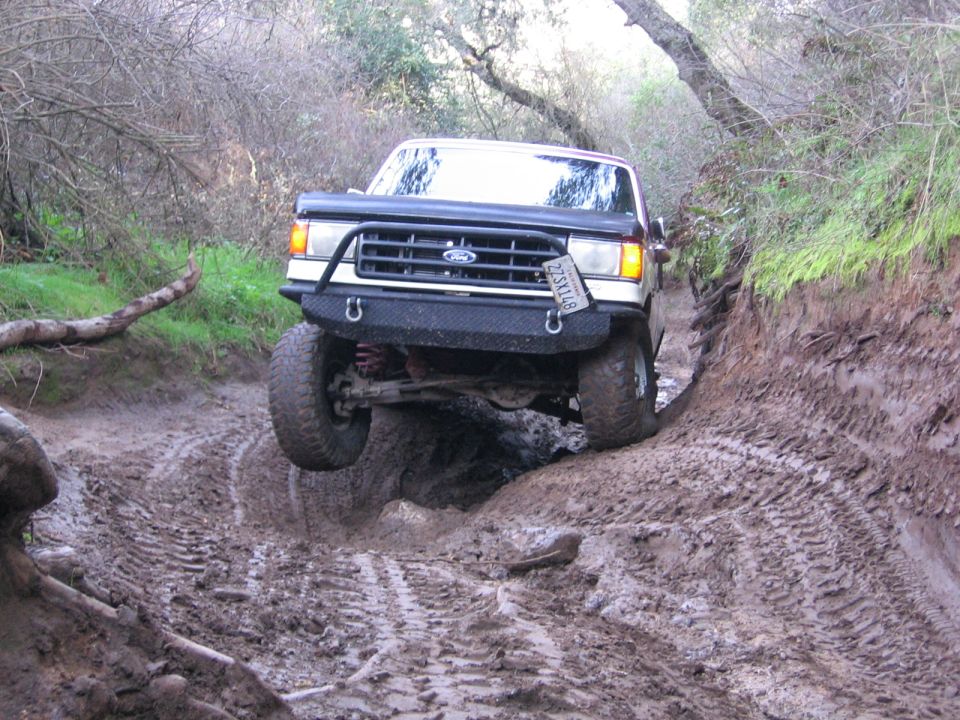 Ford Bronco with aftermarket bumper navigating a muddy rut, front wheel off the ground