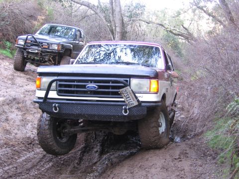 1989 Ford Bronco - Two off-road vehicles navigate a wet, muddy trail surrounded by trees and brush.