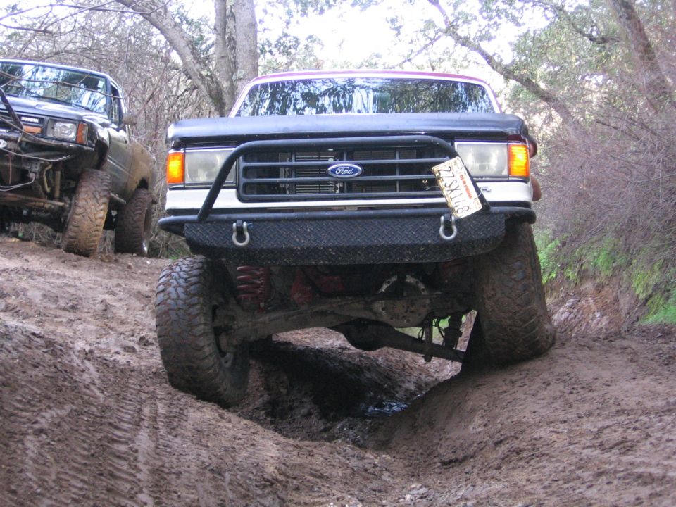Ford truck with aftermarket bumper going over an obstacle on a muddy trail. Another truck is visible to the left