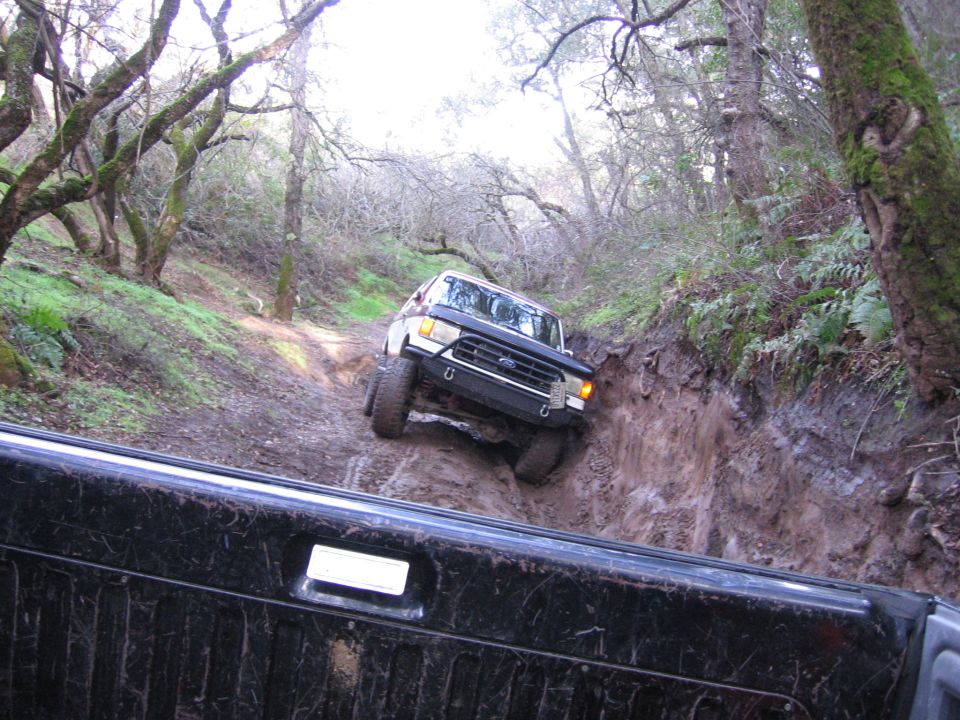 1989 Ford Bronco navigating a steep, muddy off-road trail with trees on either side