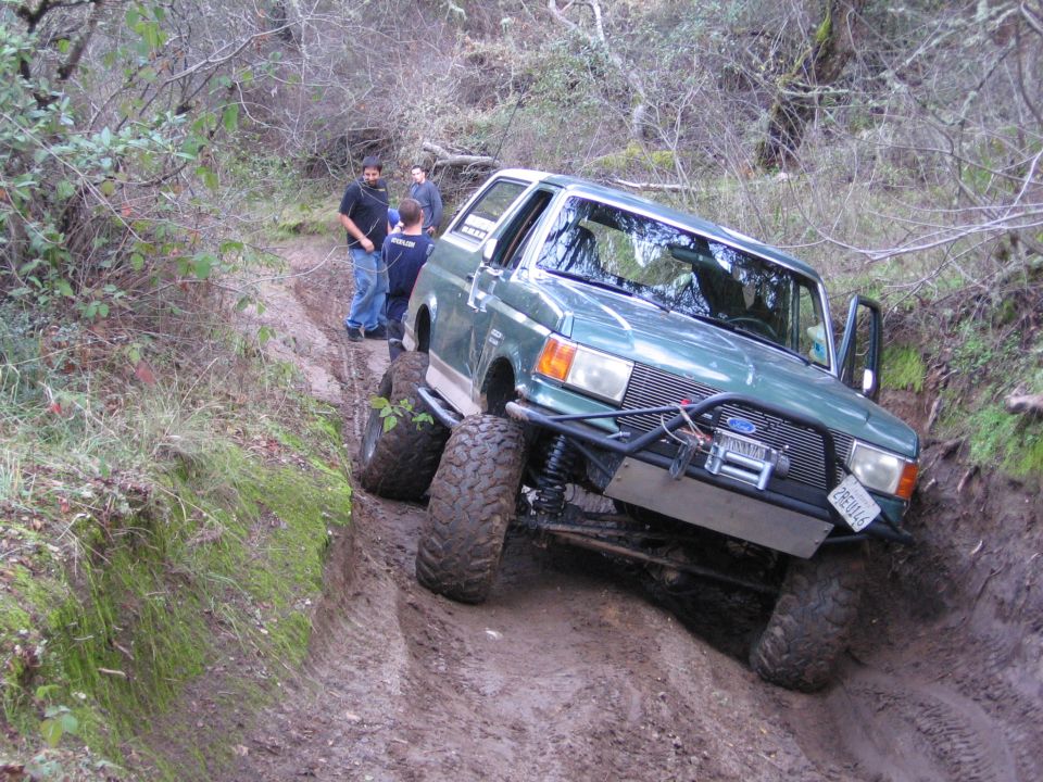 Green 1989 Ford Bronco modified for off-roading navigating a muddy trail with people watching