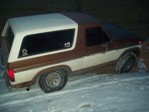 1985 Ford Bronco - A two-tone brown and white 1985 Ford Bronco is stuck in the snow, viewed from the front three-quarter angle.