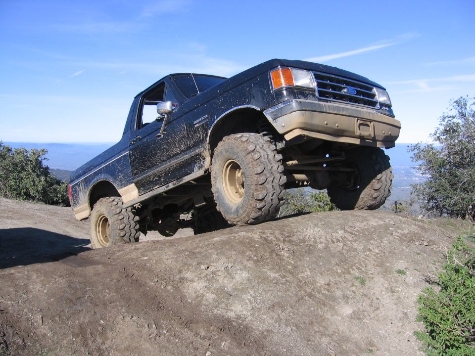 1989 Ford Bronco with large off-road tires climbing a dirt incline under a blue sky