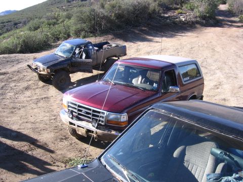 1989 Ford Bronco - Three off-road vehicles are parked on a dusty trail in a hilly, scrub-covered area.