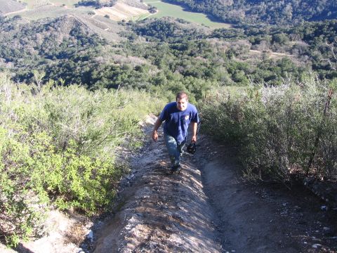 1989 Ford Bronco - A man ascends a narrow dirt trail surrounded by vegetation, with rolling hills and fields visible in the background.