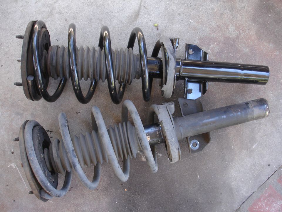 Two car suspension struts, one black and one gray, laid on a concrete surface