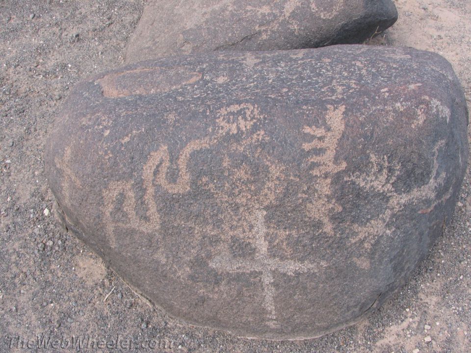 Dark boulder with petroglyphs of figures and symbols, set in gravelly terrain