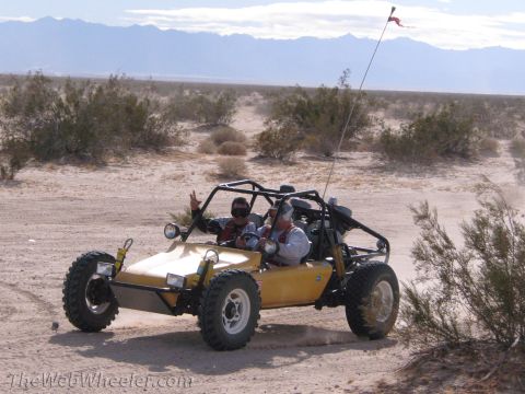 1996 Ford C-350 (Centurion Conversions) - Two people ride in a yellow dune buggy through a desert landscape with mountains in the background.