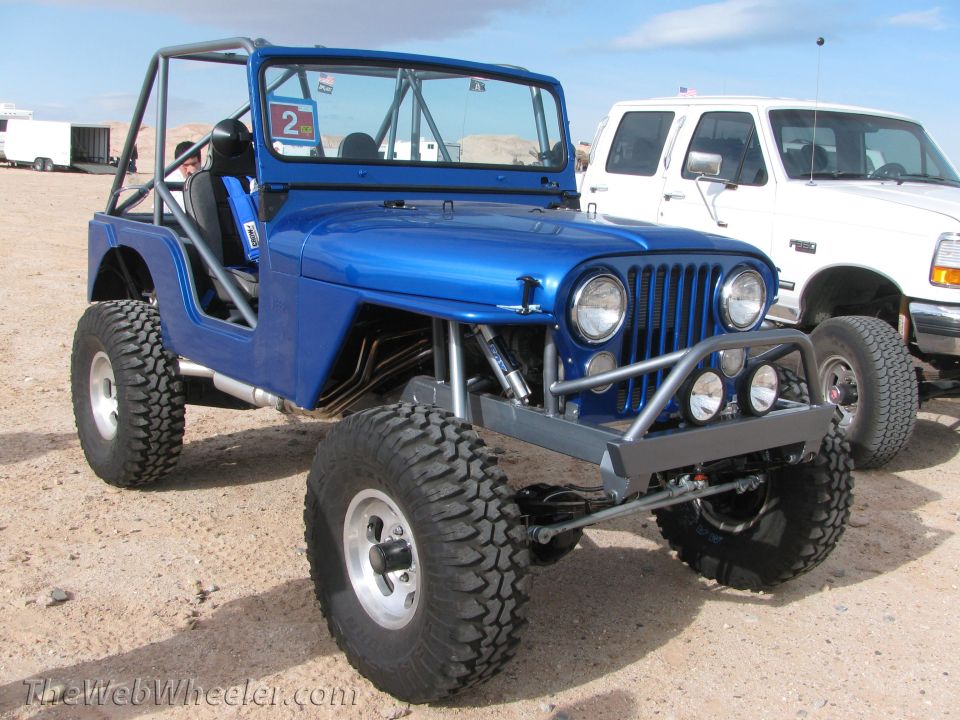 Blue off-road Jeep with roll cage and large tires parked next to a white truck