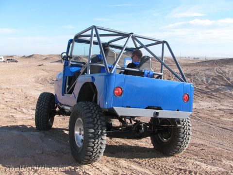 1996 Ford C-350 (Centurion Conversions) - A bright blue, modified jeep with a prominent roll cage is shown from the rear, parked on a sandy terrain under a clear sky.