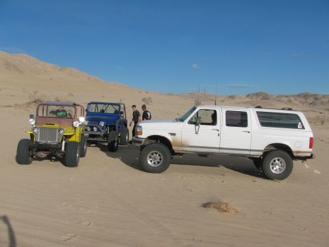1996 Ford C-350 (Centurion Conversions) - Three off-road vehicles, including a white pickup truck and two Jeeps, are parked in a sandy desert landscape under a clear blue sky.