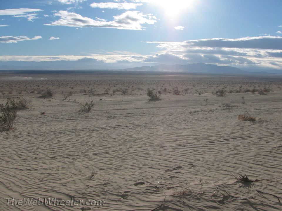 Vast desert with sand dunes, sparse brush, and mountains in the distance under a bright sun and cloudy sky