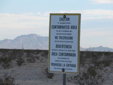 1996 Ford C-350 (Centurion Conversions) - A bilingual white-and-yellow warning sign on a steel post in open desert scrub reads 'CAUTION U.S. GOVERNMENT PROPERTY / CONTAMINATED AREA…