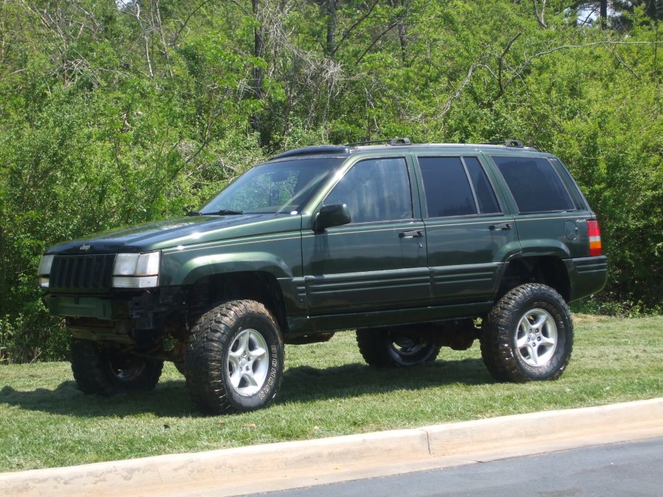 Dark green lifted Jeep Grand Cherokee with big off-road tires parked on grass.
