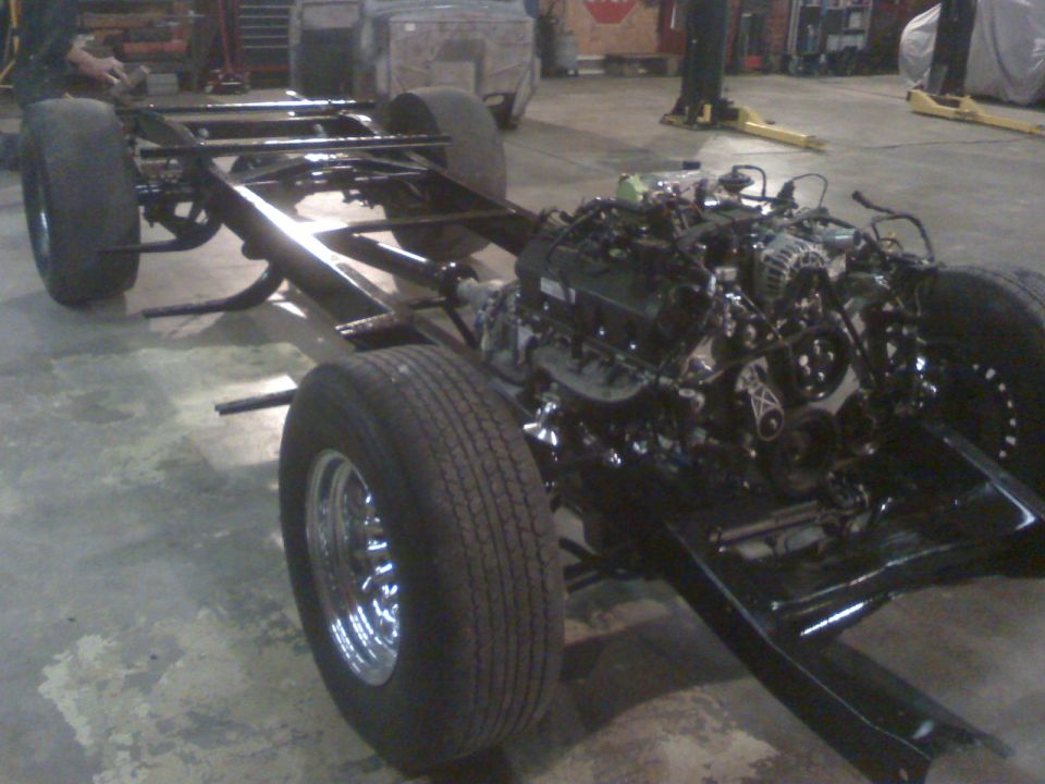 Black rolling chassis with V8 engine and chrome wheels in a shop, no body mounted