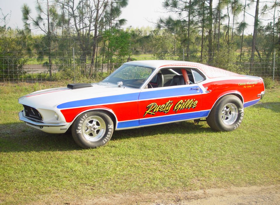 White and red 1969 Ford Mustang drag car with blue stripe and 'Rusty Gillis' text, parked on grass