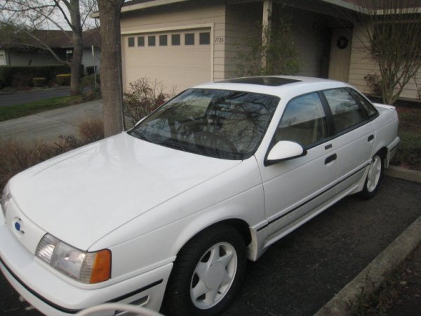 White sedan parked in driveway, 3/4 front view, showing front bumper, hood, driver's side fender, door, and rear quarter panel