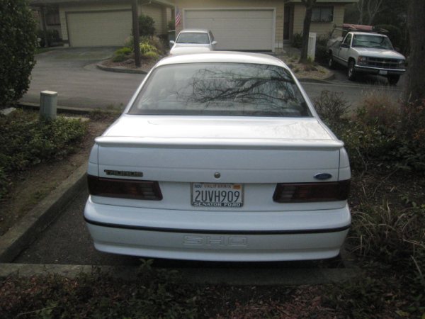 Rear view of a white 1993 Ford Taurus parked in a driveway. Another car is in the garage behind it, and a pickup truck is parked to the right