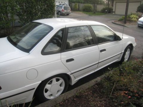 1993 Ford Taurus - A white sedan is parked on the side of the road, showing its profile with black trim and distinctive rear quarter window.