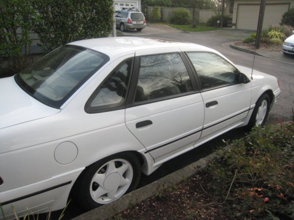 White sedan parked on a curb, side view. The car has black trim and a triangular rear quarter window