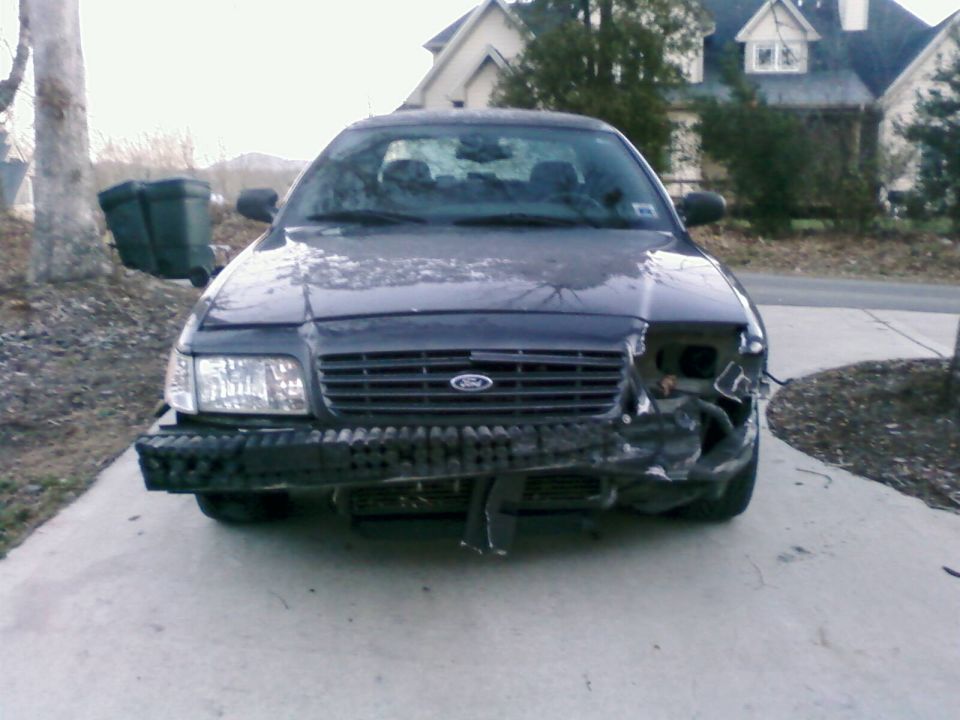 Front view of a dark-colored Ford Crown Victoria with significant front-end damage, missing headlight and bumper