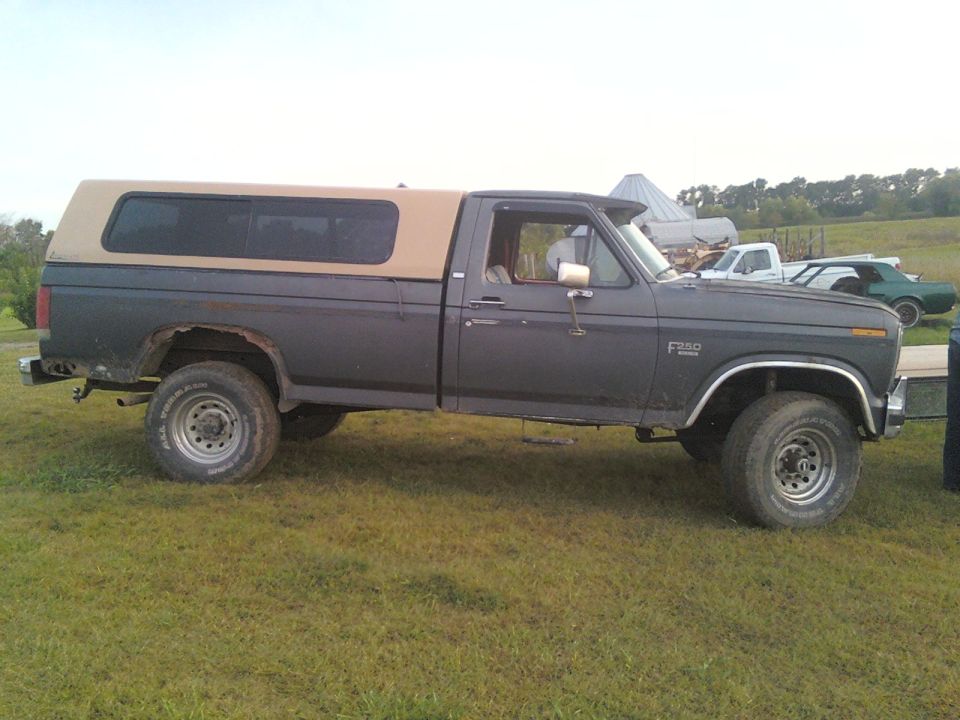 Ford F-250 pickup truck with a tan camper shell, parked in a grassy field. Other vehicles are visible in the background