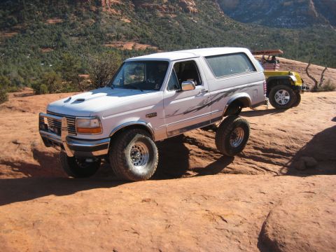 1996 Ford Bronco - A lifted white Ford Bronco navigates rocky terrain, with a yellow Jeep in the background.